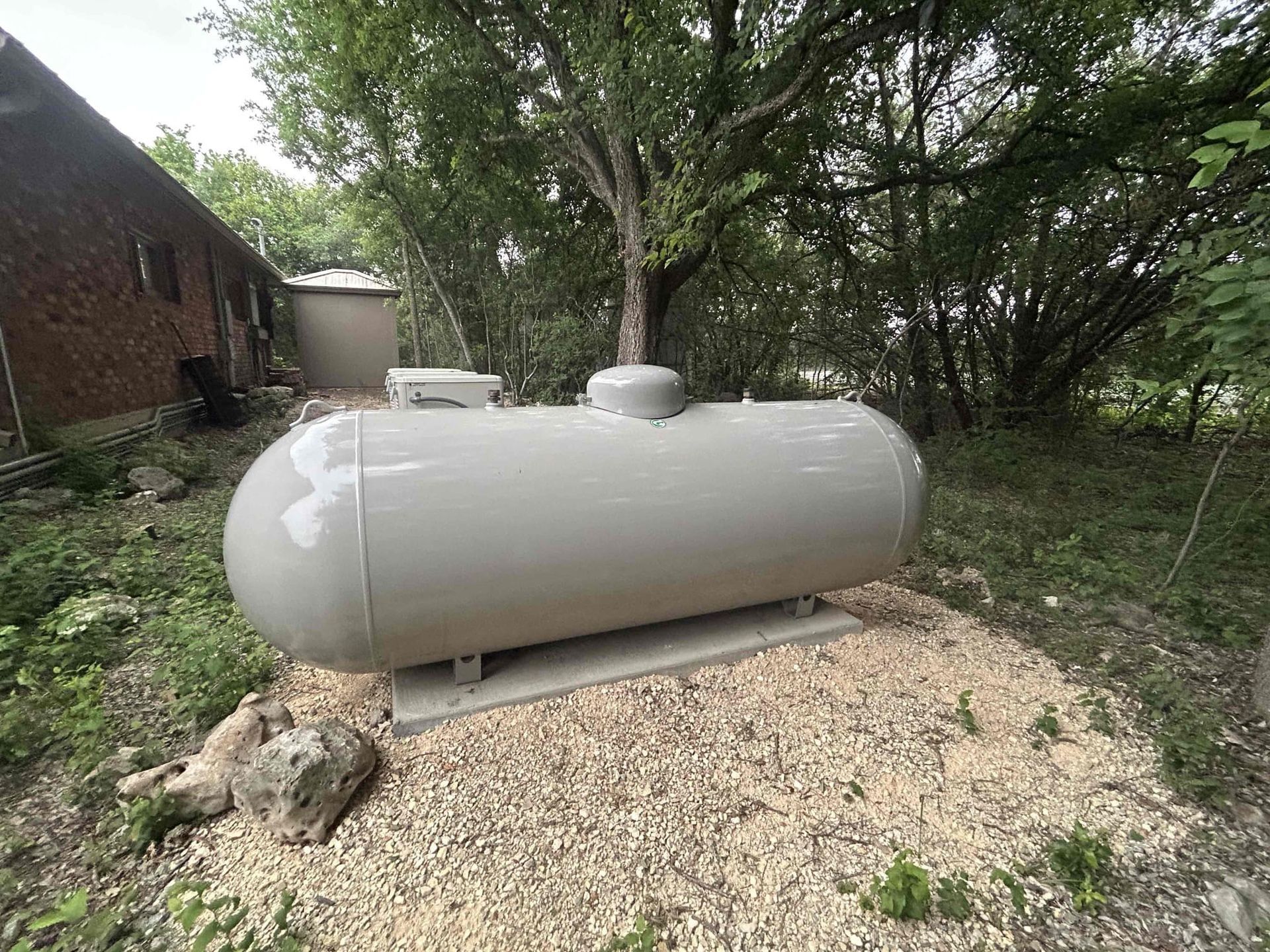 Gray propane tank on a gravel mound beside a brick building and trees outdoors