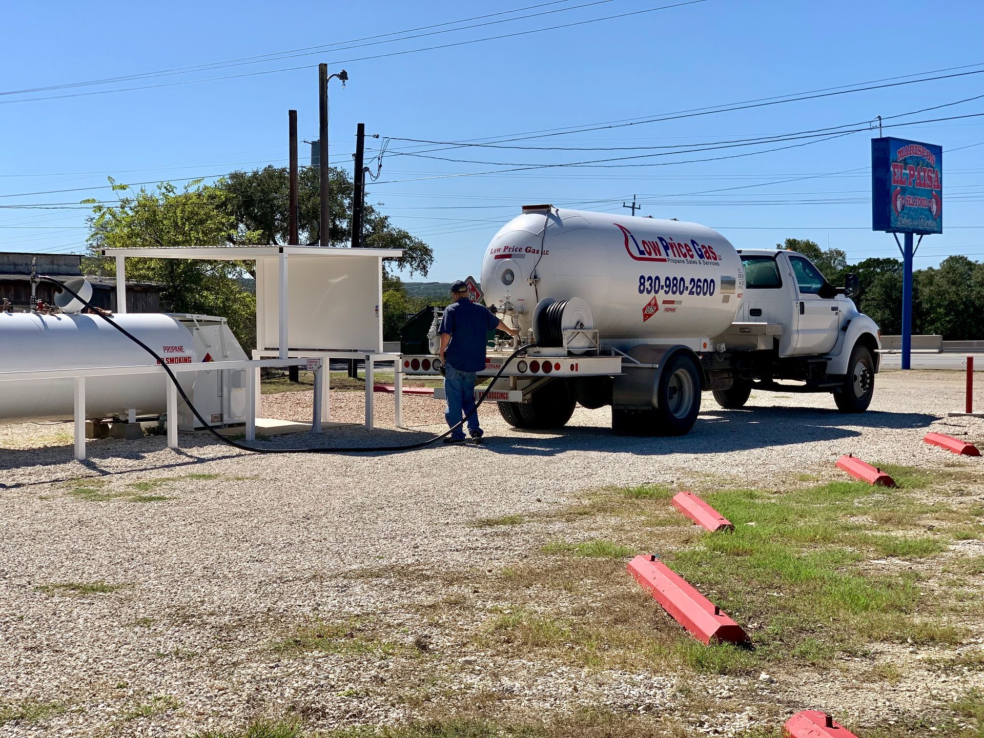 Worker beside white tanker truck at a roadside fuel station on a sunny day.