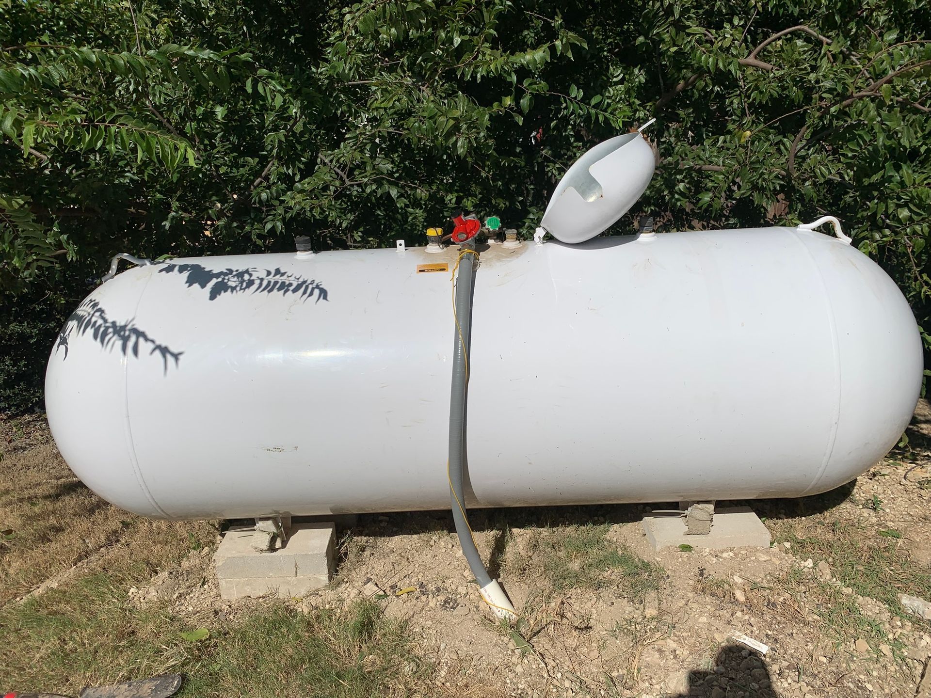 White propane tank on concrete blocks outdoors with a valve and hose against a leafy background