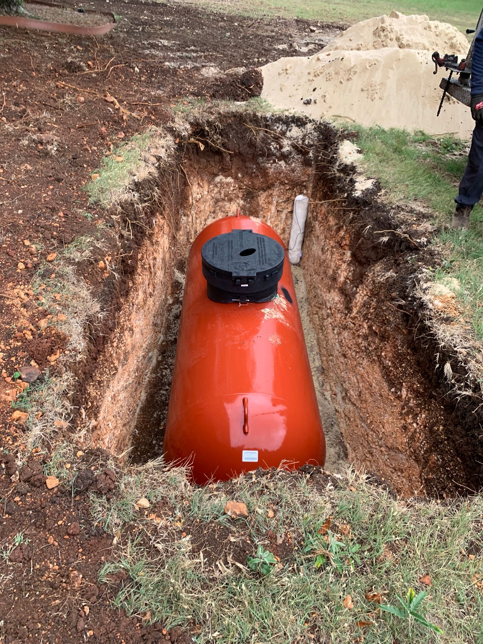 Orange water tank buried in a trench outdoors, with a black cap on top.