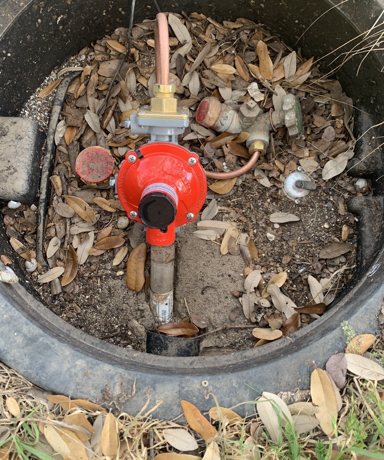 Red water meter/valve inside a round concrete utility box filled with dirt and leaves