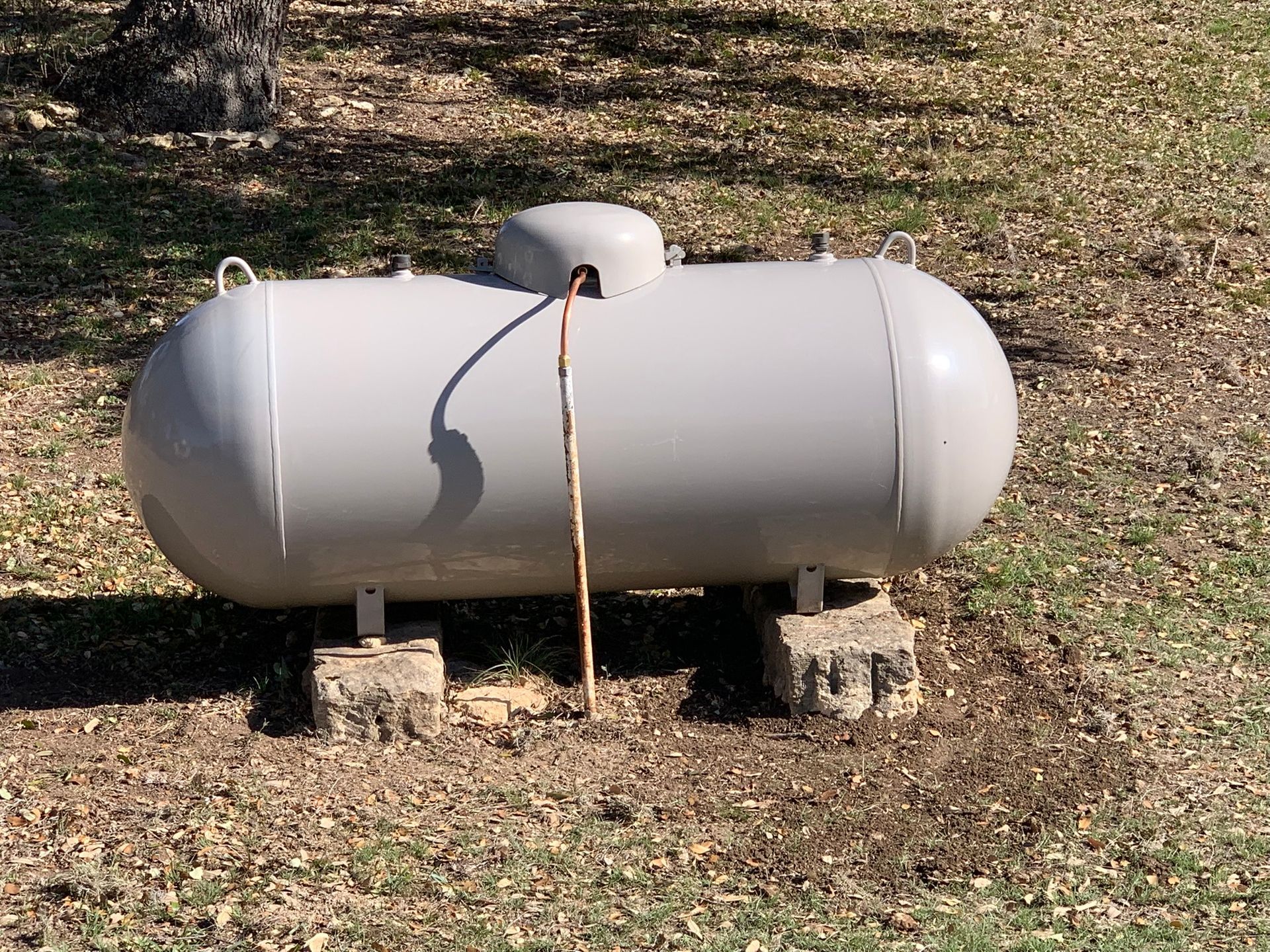 Gray propane tank on blocks in a grassy yard, with a hose attached.