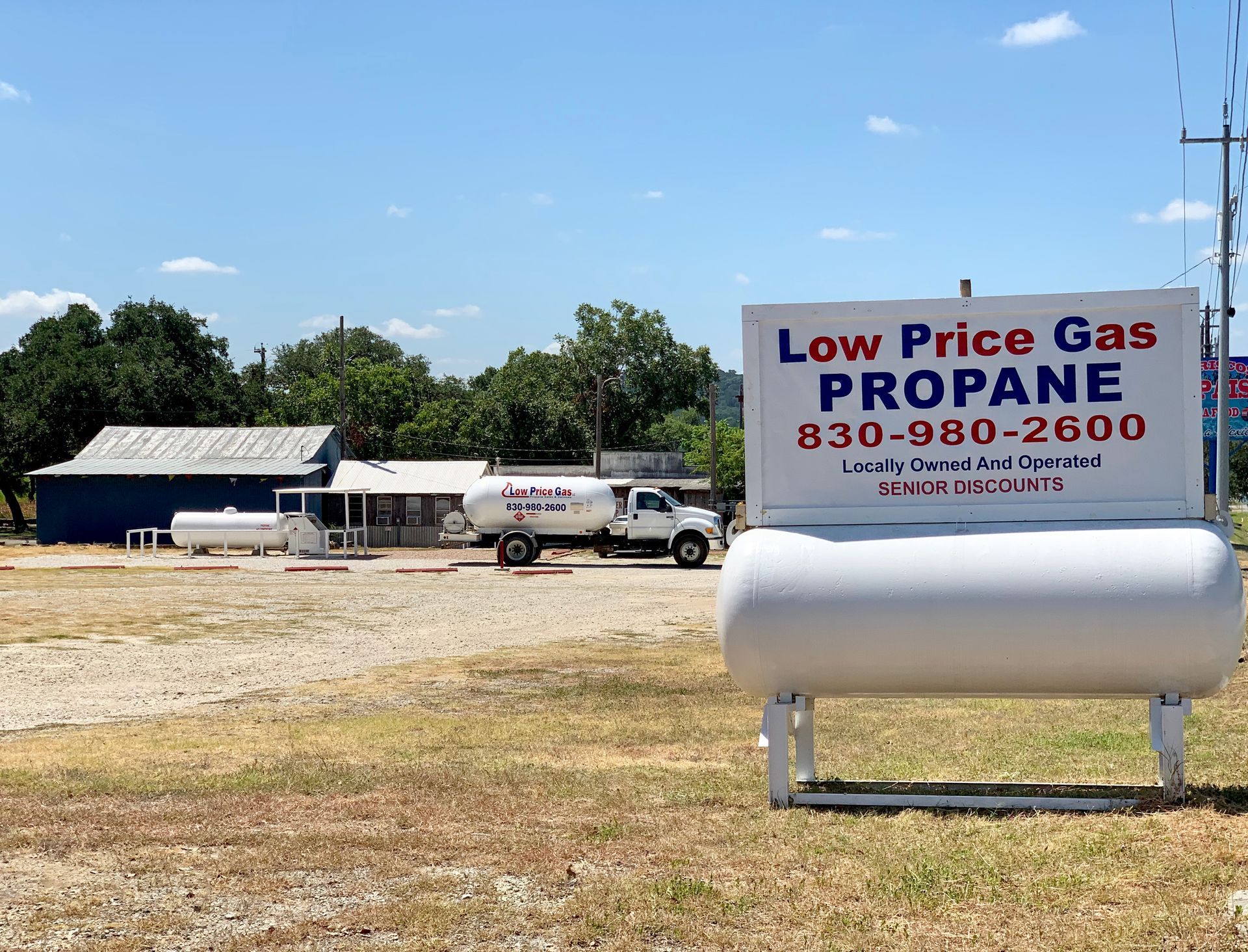 Low Price Gas propane tank and delivery truck beside a rural roadside sign under a blue sky