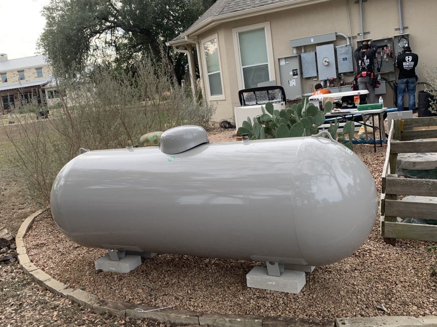 Gray propane tank beside a house on gravel, with utility equipment in the background