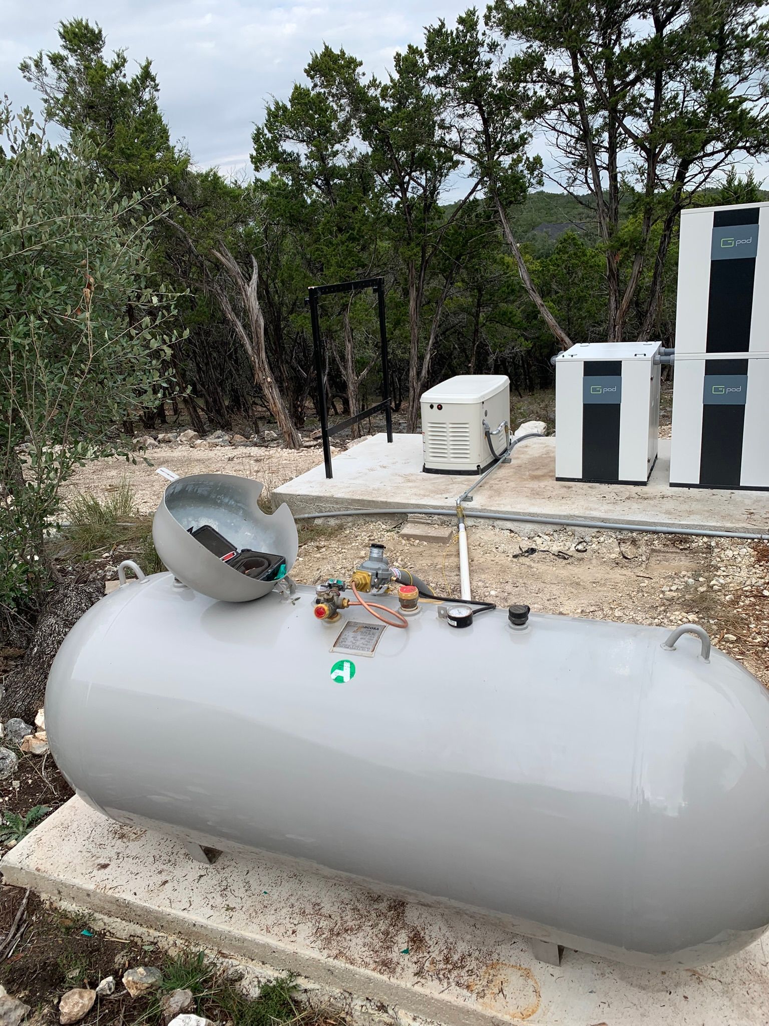 Horizontal gray propane tank outdoors with gauge and valves near white utility boxes and trees