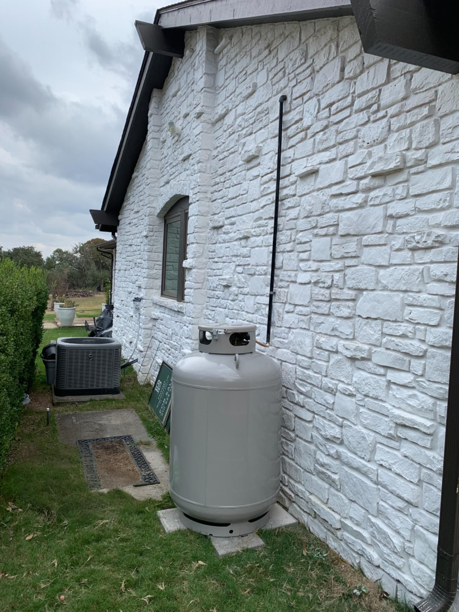 Gray propane tank beside a white stone house with grass and cloudy sky nearby