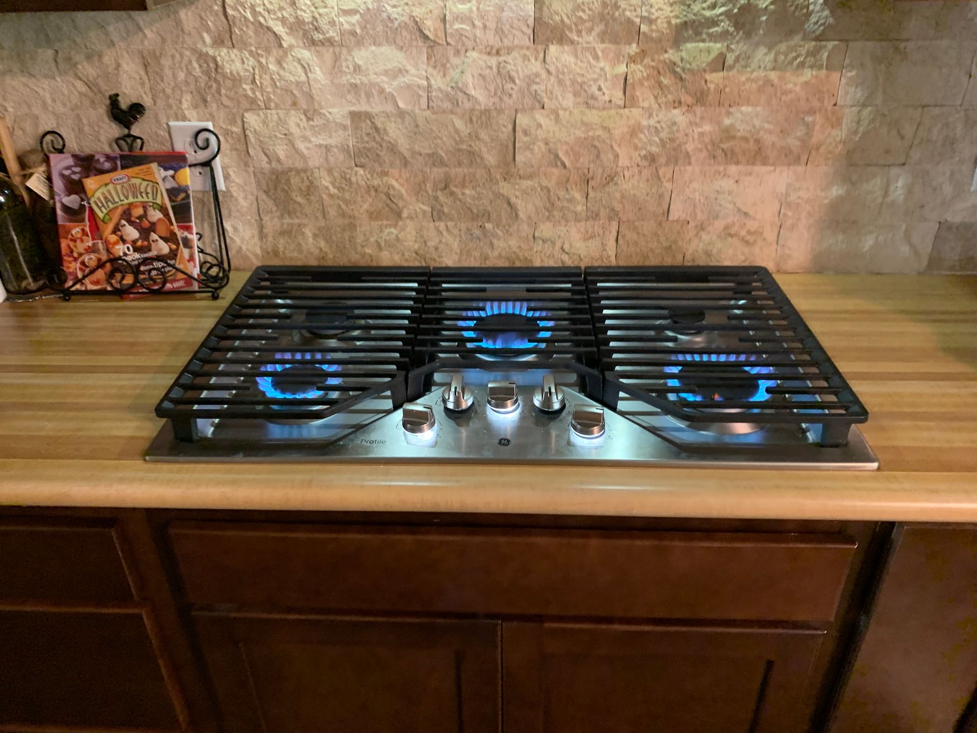 Black gas cooktop with three blue flames on a wooden kitchen counter against a stone backsplash