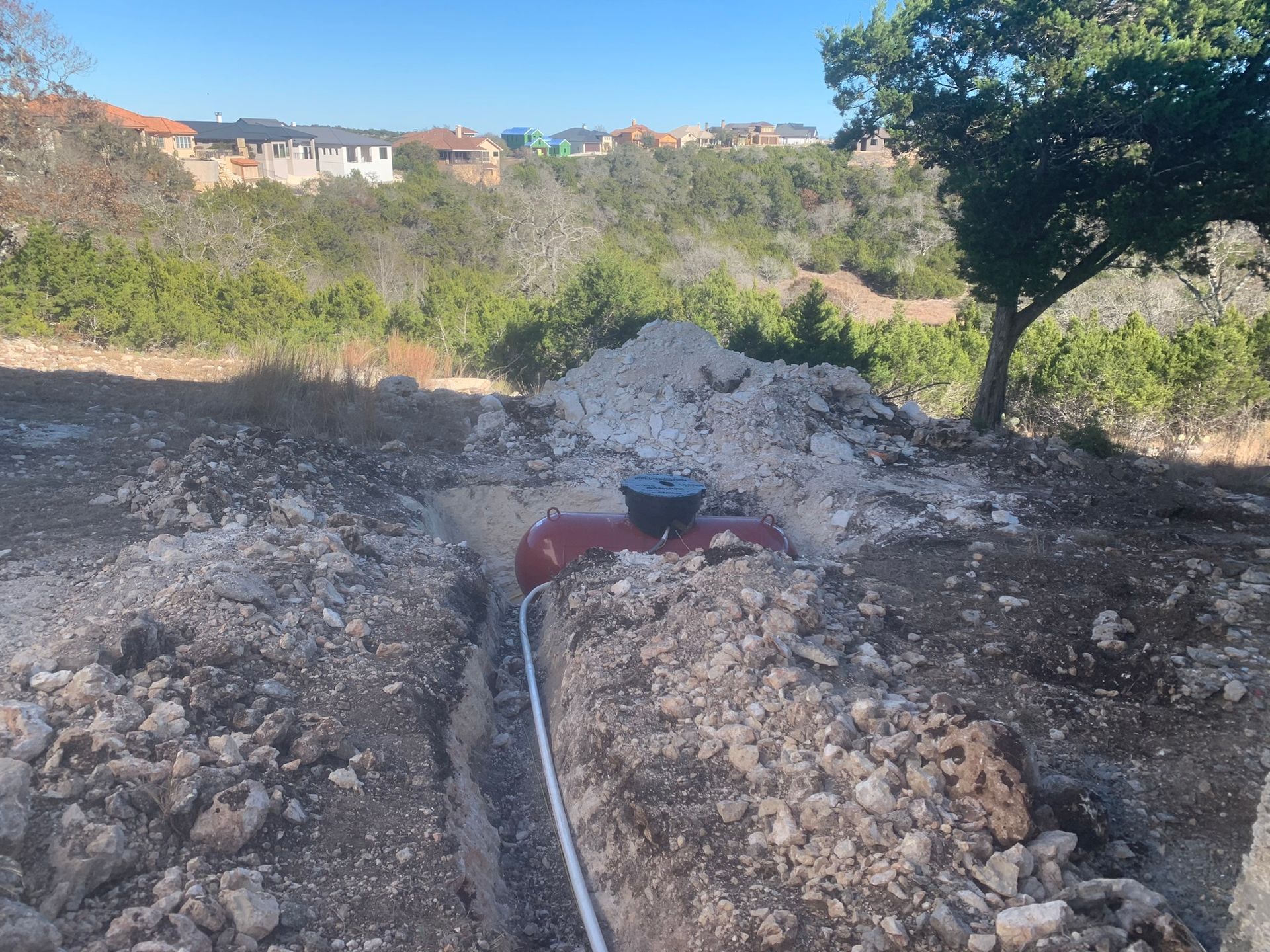 A red wheelbarrow on a rocky dirt path beside a trench, with trees and houses in the background.