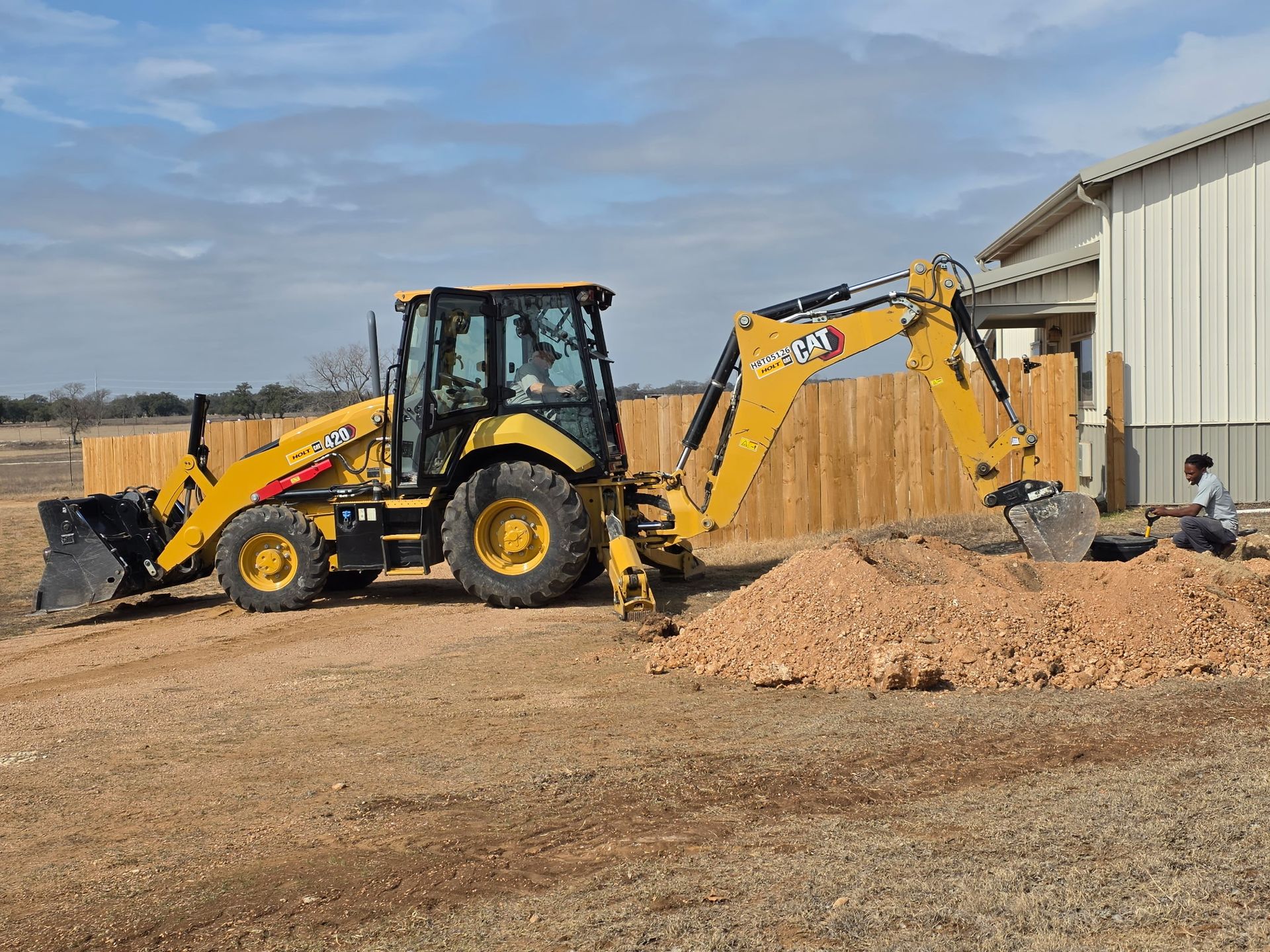 Yellow backhoe loader parked beside a metal building, scooping wood chips in a gravel lot.