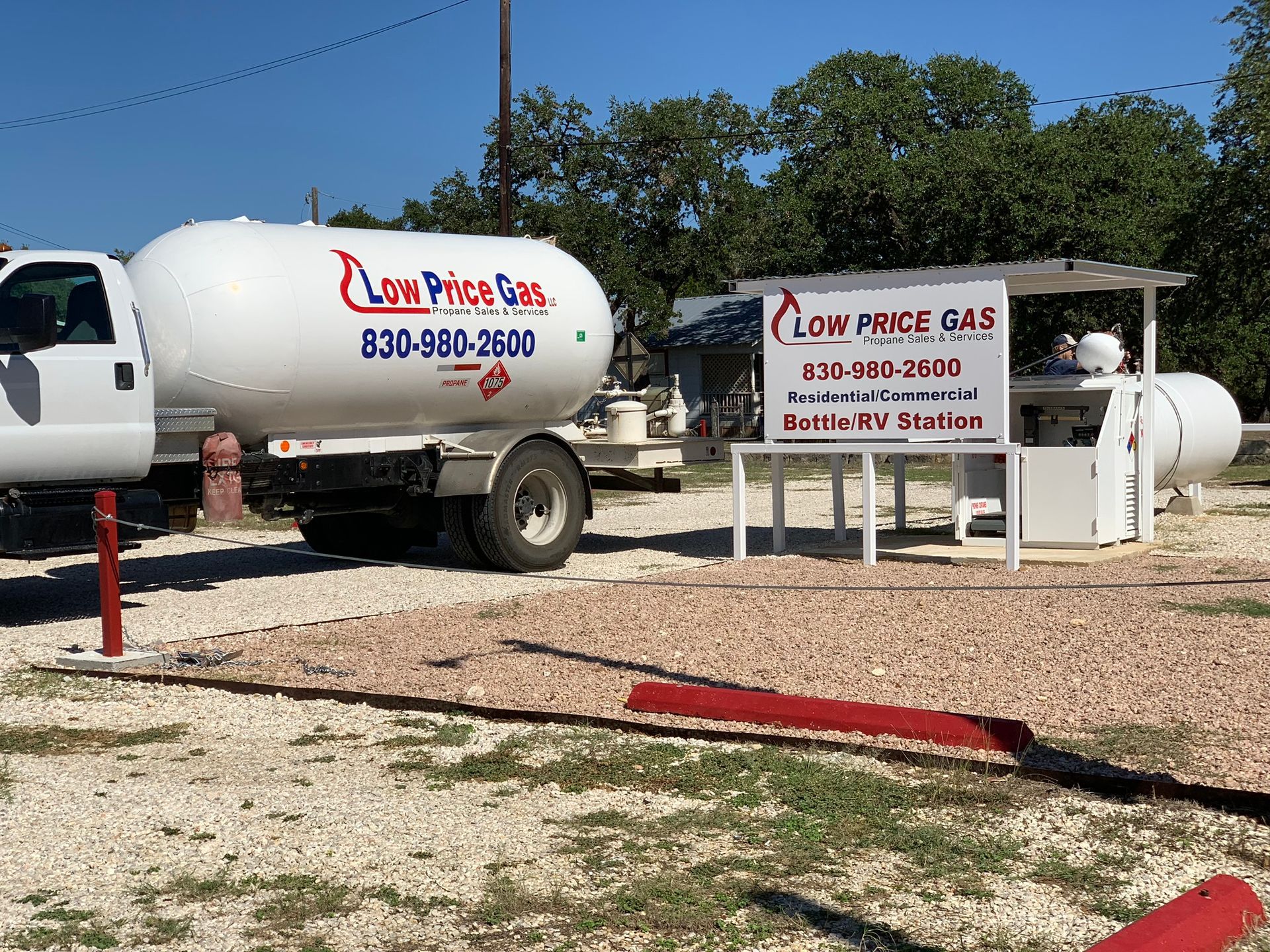 White propane truck and tank with “Low Price Gas” sign on a gravel lot under trees.