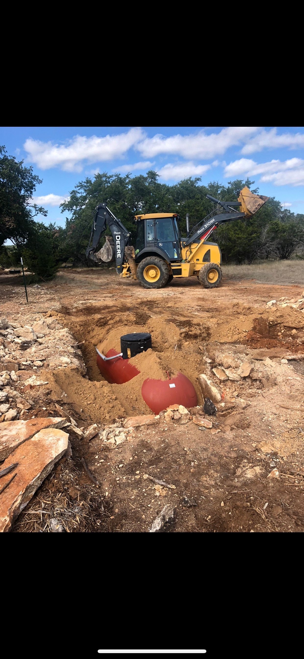 Yellow backhoe at a dirt excavation site with two red barrels in the foreground and trees behind.