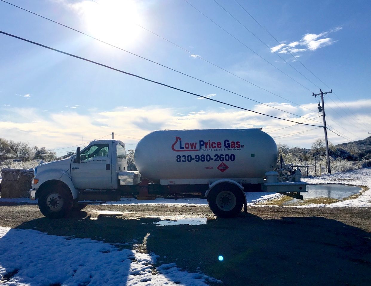 White propane delivery truck with tank marked “Low Price Gas” parked on a snowy roadside under a bright sky