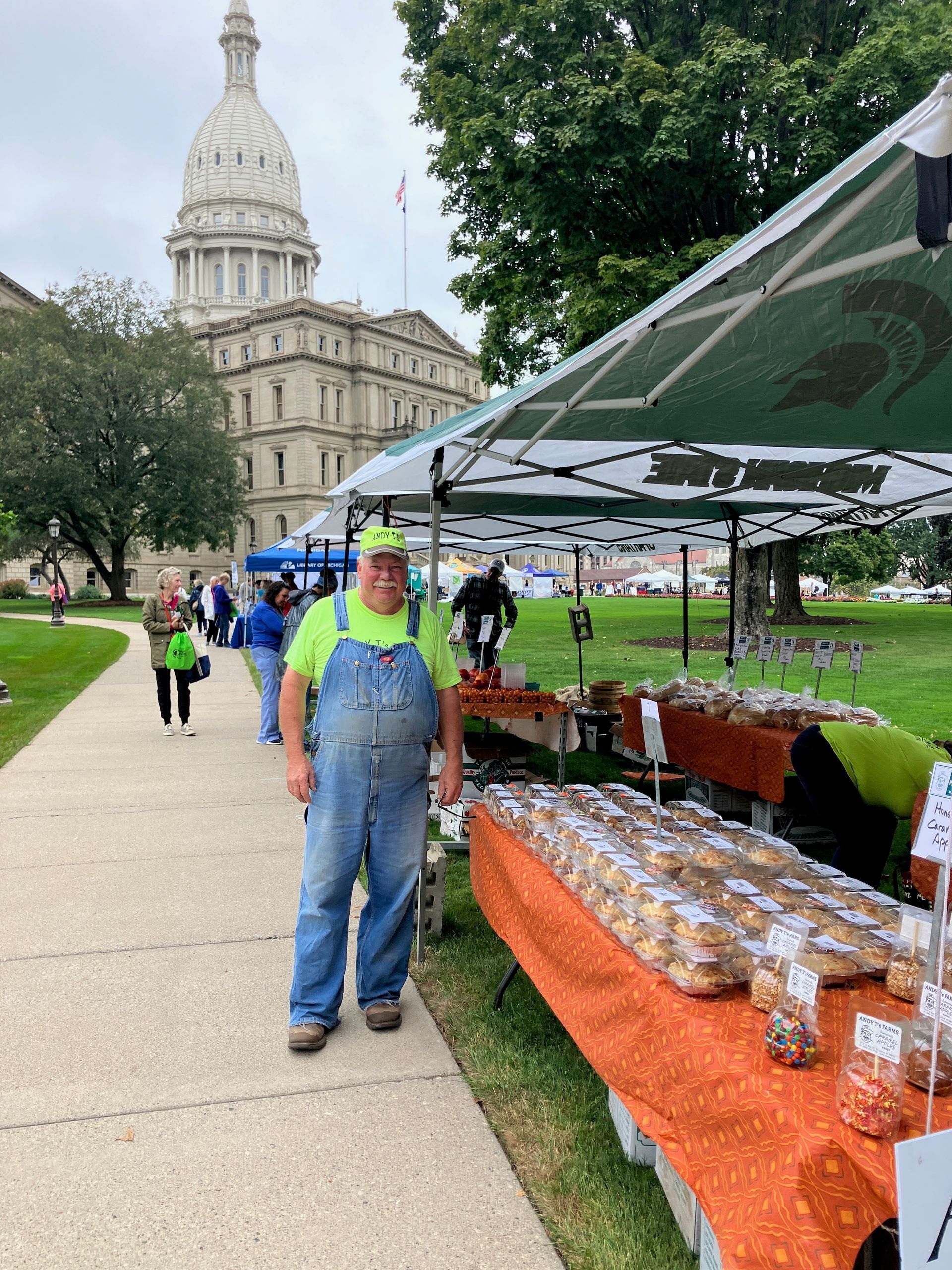 A man in overalls is standing in front of a food stand.