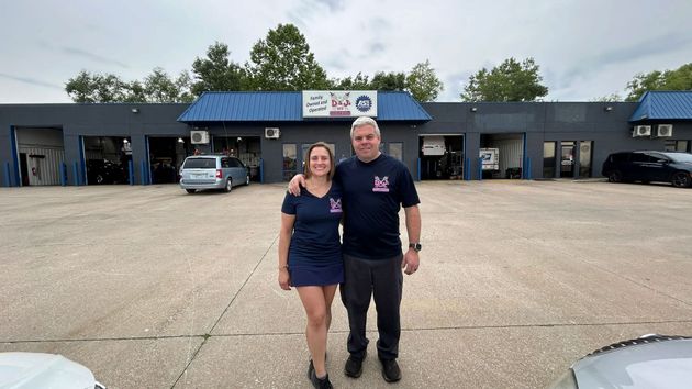 Two people standing in a parking lot in front of a low building with a blue roof and open garage bays