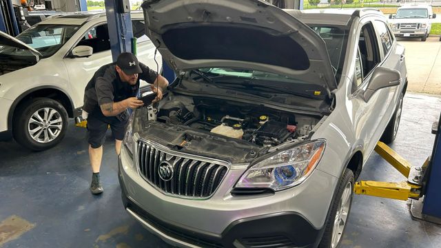 Man using a tool beside a silver SUV with hood open, inspecting the engine in a parking garage