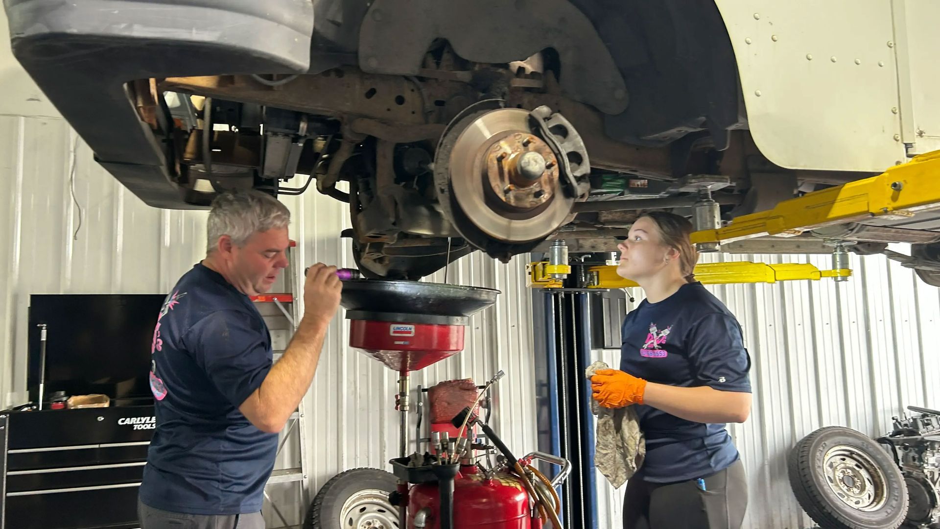 Two mechanics under a raised car, inspecting the exposed wheel hub in an auto shop