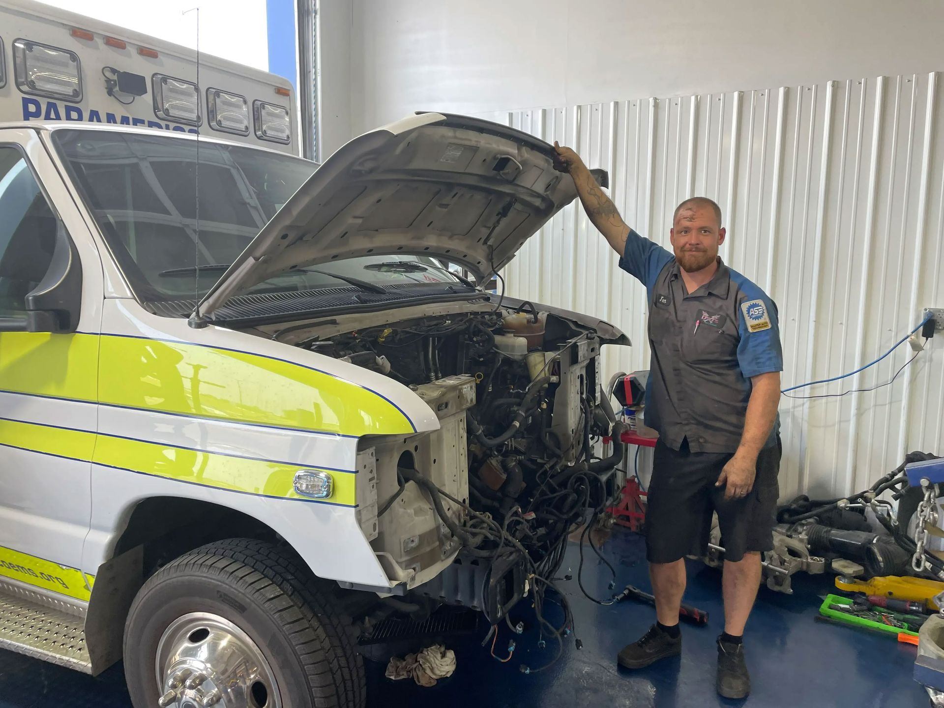 Mechanic stands beside open-hood ambulance in garage, inspecting exposed engine under fluorescent lights.