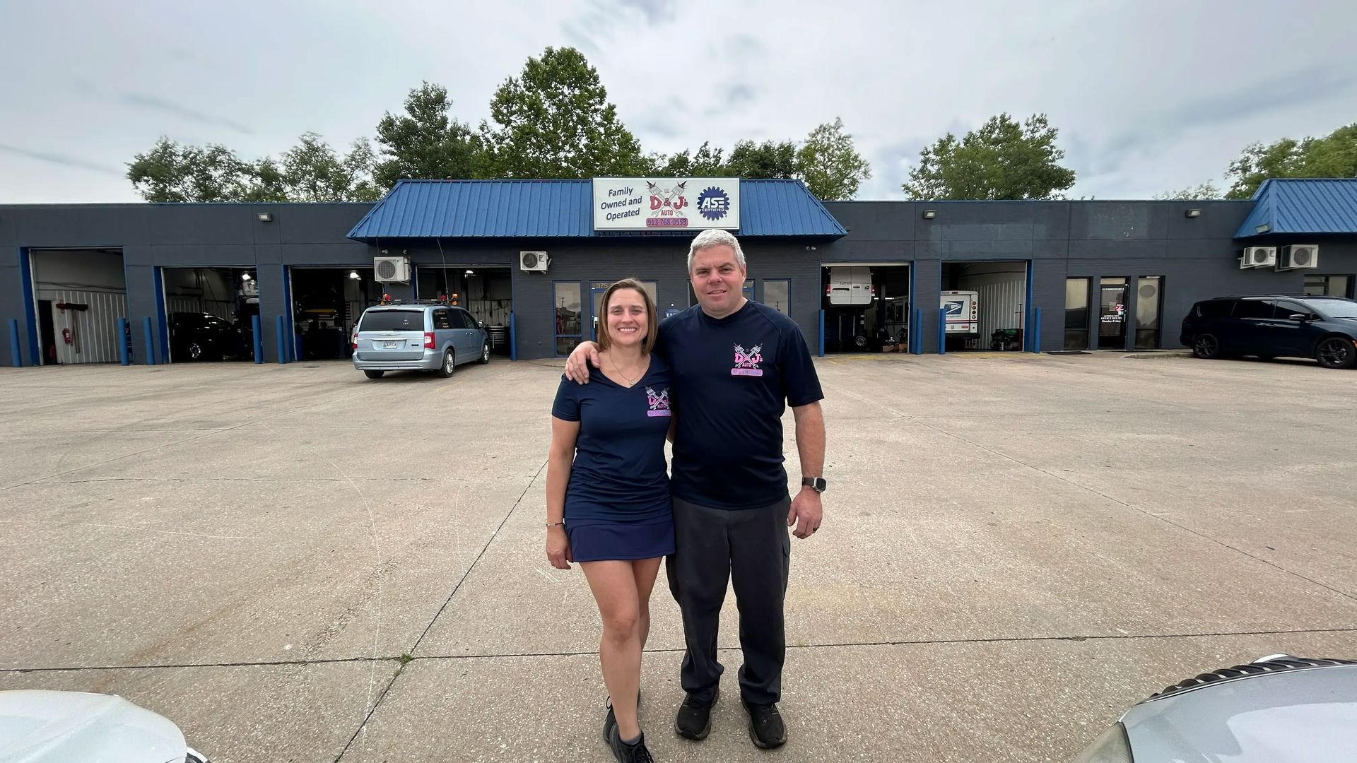 Two people standing in front of a blue-roofed business building with parked cars and open garage bays