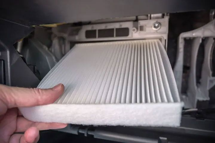 Hand holding a white pleated HVAC cabin air filter in front of a car filter slot