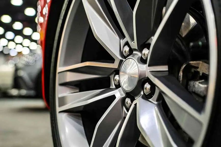 Close-up of a silver alloy car wheel on a vehicle in a dim garage, with blurred lights in the background