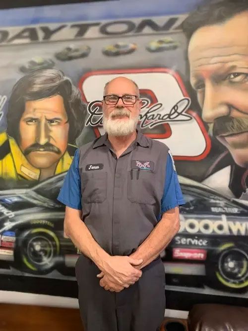 Man standing in front of a racing mural with cars and portraits, wearing a gray work shirt.