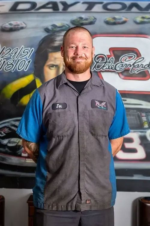 Man standing in front of a race car mural, wearing a gray and blue work vest, smiling.