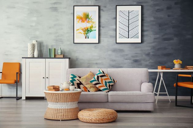Modern living room with light gray sofa, woven poufs, orange chair, and framed art on a concrete wall