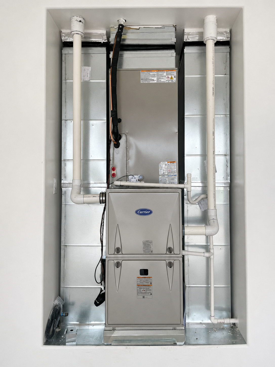 Furnace and metal ductwork installed in a utility closet with exposed piping and wiring.