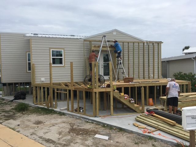Workers building a wooden deck on a beige mobile home under a cloudy sky