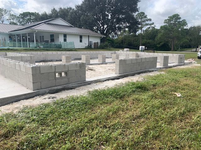 Partially built concrete foundation with walls in a grassy yard near a white house and trees