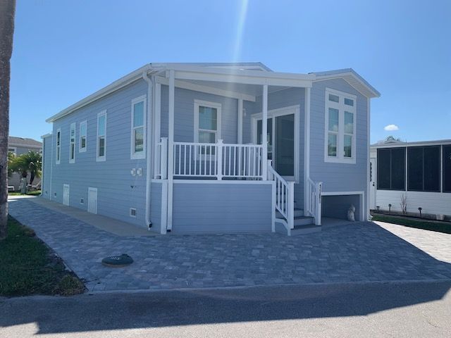 Light blue house with white trim and a small porch on a blue brick driveway, sunny day.