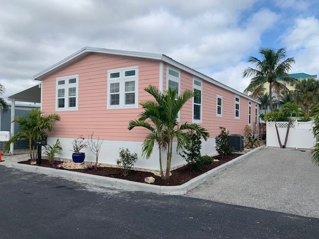 Pink mobile home with white trim and a palm tree in the front yard. Driveway. Cloudy sky.
