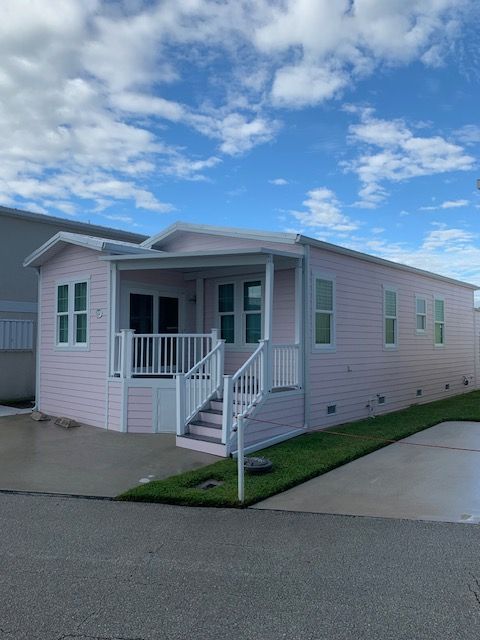 Small pink manufactured home with white trim, front porch, and steps beside a paved street under a blue sky