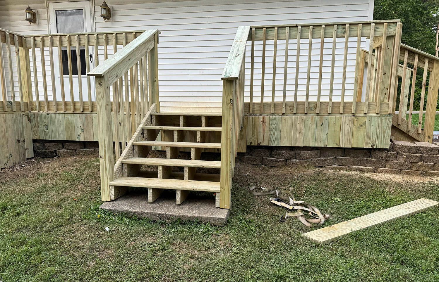 Wooden front steps and ramp on a small porch beside a white house, with railing and grass below.