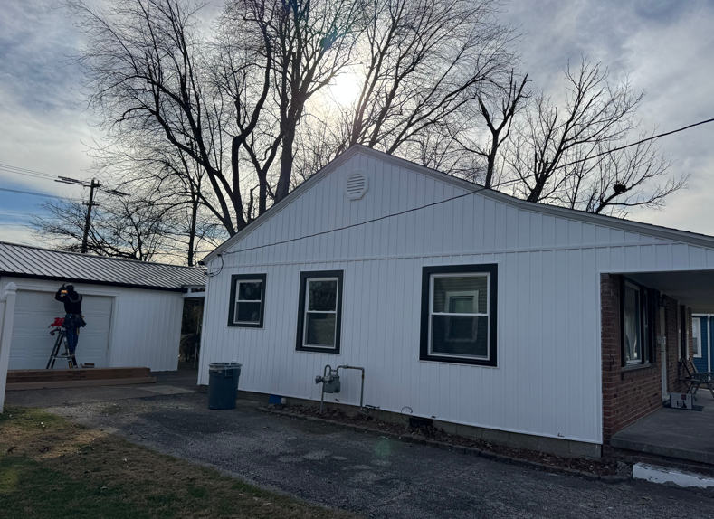 White house with black-trimmed windows and bare trees; a person is climbing a ladder at the left garage.
