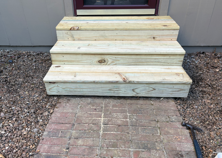 Wooden steps leading to a doorway, with a brick walkway and gravel on either side.