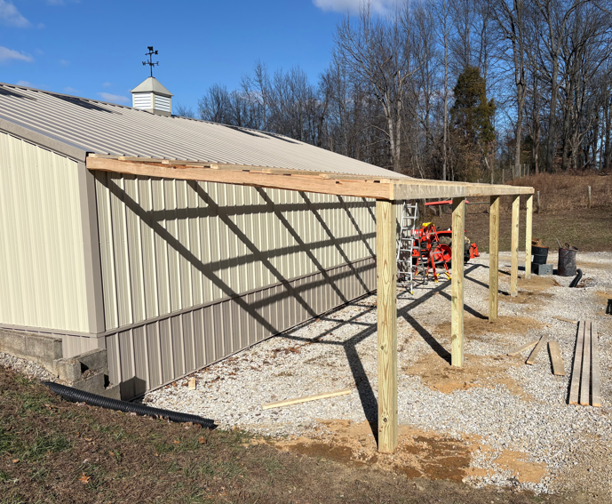 Partially built wooden carport beside a beige barn on a gravel lot, with construction materials nearby