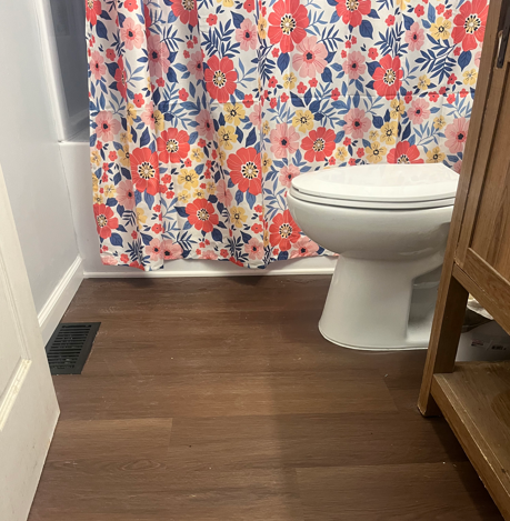 Small bathroom with white toilet, floral shower curtain, wood floor, and vent near the tub.