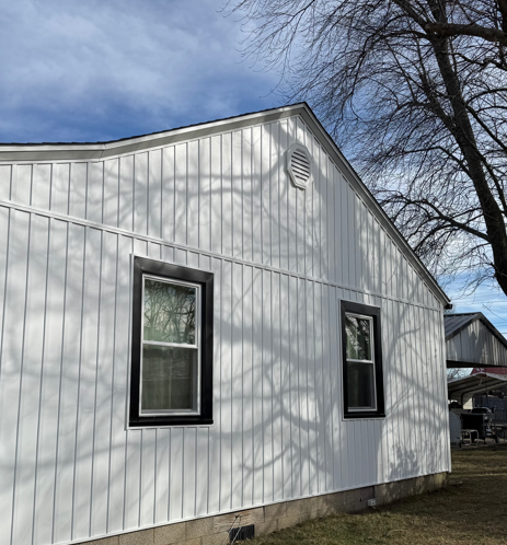 White house siding with two windows and a vent under a blue sky with tree shadows