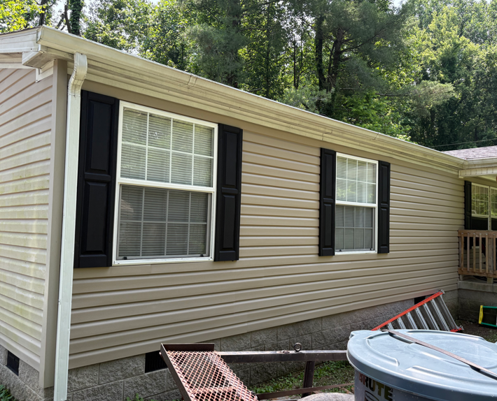 Tan-sided house exterior with black shutters and windows beside a wooded yard