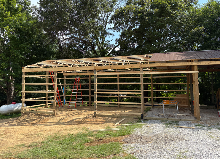 Partially built wooden carport frame with ladders, surrounded by trees and gravel driveway