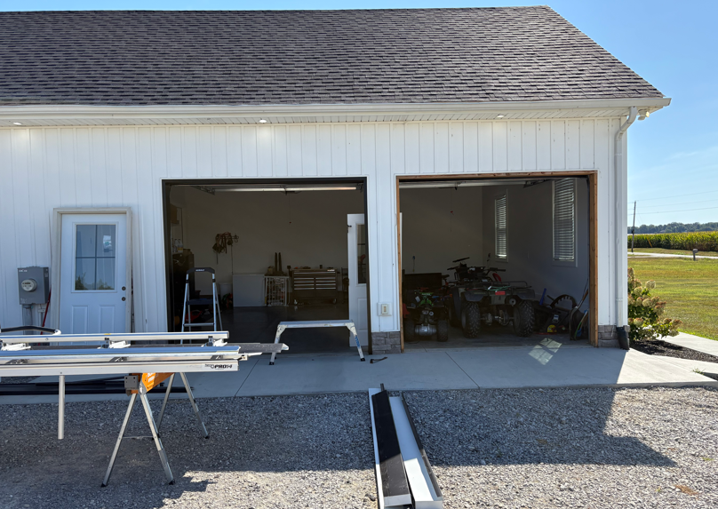 White garage with two open bays, workbench, bicycles, and a bench in front on a gravel driveway