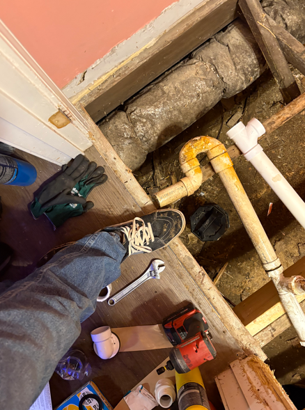 Person standing in a floor opening with exposed pipes and tools, inspecting plumbing work.