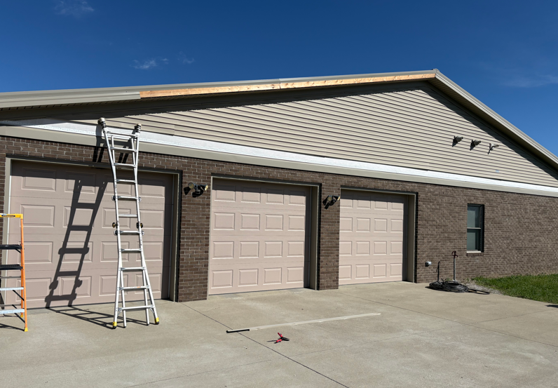 Residential garage with beige doors, brick exterior, and two ladders leaning against the roofline.
