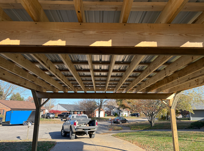 Under a wooden carport, a pickup truck is parked in a suburban driveway on a sunny day.