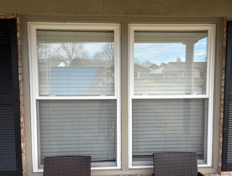 Two white-trimmed windows with blinds on a beige exterior wall, with two chair backs below.