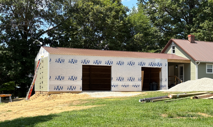 New house under construction with white siding, garage opening, and piles of lumber on a grassy lot
