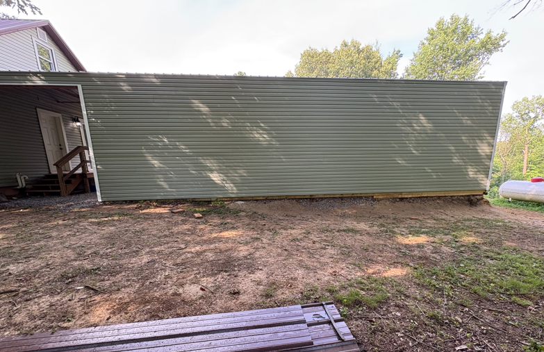 Side view of a long green shed or outbuilding beside a house, with bare dirt in front.