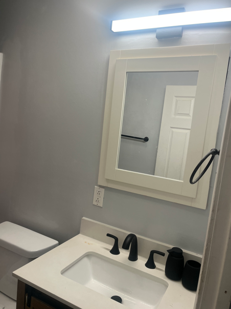 Bathroom vanity with mirror, light, black faucet, and towel ring beside a toilet.