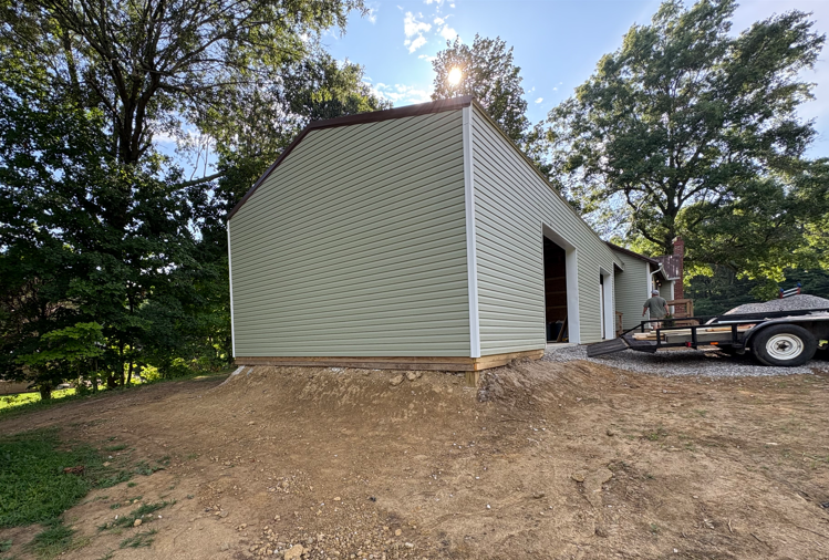 Side view of a light green detached garage with a dirt yard and a truck parked beside it.